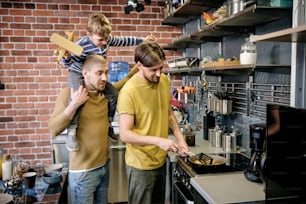 Young gay man cooking breakfast on stove in kitchen when his husband giving piggyback ride to their little son