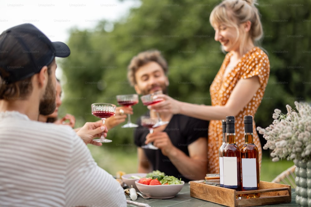 Os jovens amigos têm um almoço festivo ao ar livre, brindando e bebendo vinho, passando um feliz verão juntos. Concentre-se em uma garrafa com lábios em branco para copiar e colar na mesa