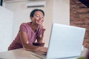 Online. Young mulatta in the kitchen watching something on internet and smiling