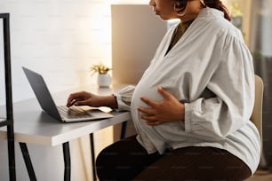 Cropped side view portrait of pregnant African-American woman using laptop at home, focus on big belly