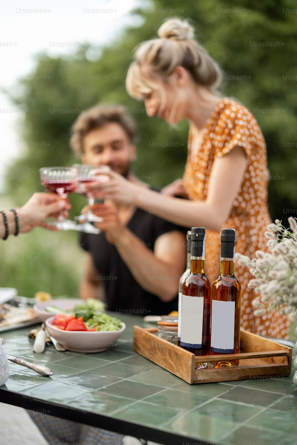 若い友達は屋外でお祝いのランチをし、乾杯してワインを飲み、幸せな夏の時間を一緒に過ごします。空白のラベルが付いたボトルに焦点を合わせて、テーブルに貼り付けます