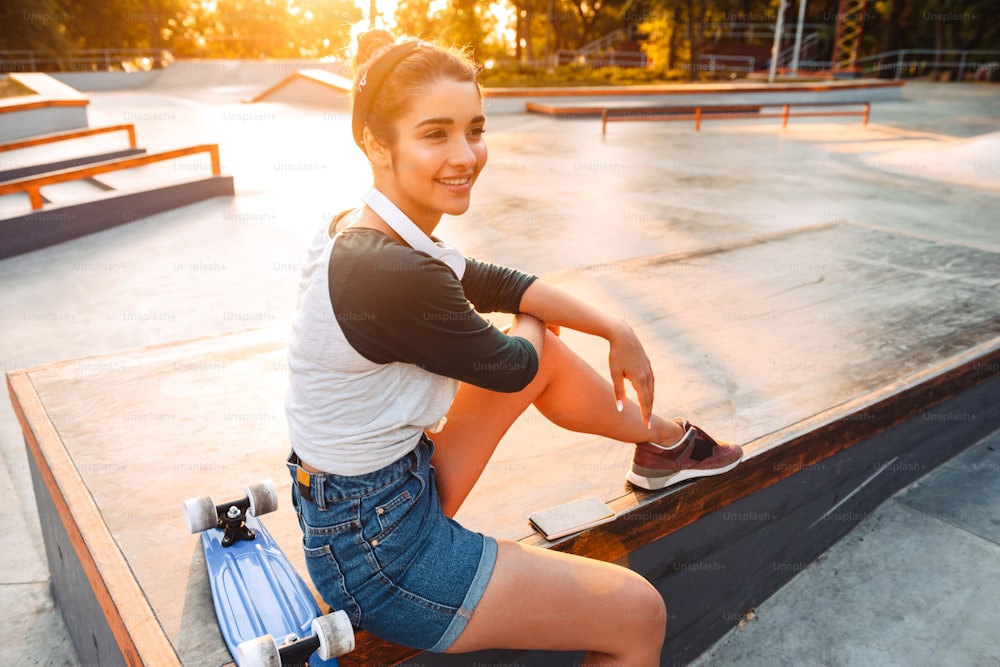 Ragazza felice con le cuffie e lo skateboard che si siede al parco