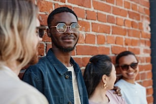 Diverse group of contemporary young people posing outdoors while standing by brick wall in urban setting, focus on smiling African-American man wearing sunglasses
