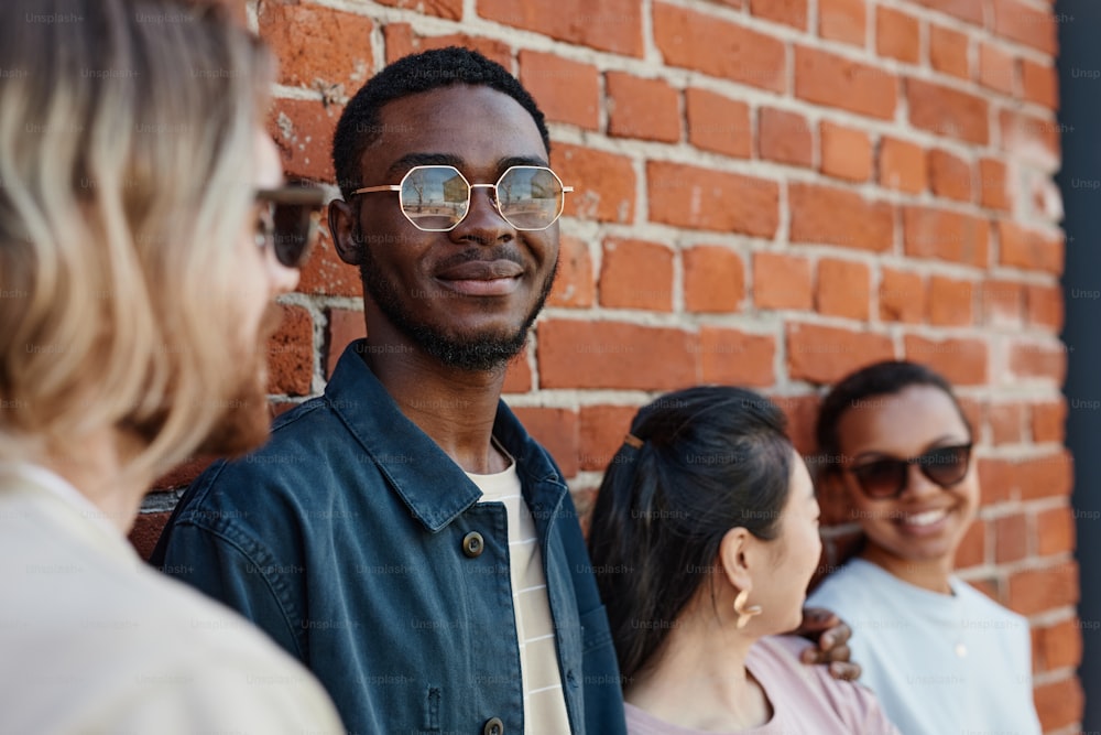 Un grupo diverso de jóvenes contemporáneos posando al aire libre mientras están parados junto a una pared de ladrillo en un entorno urbano, se centran en el hombre afroamericano sonriente con gafas de sol