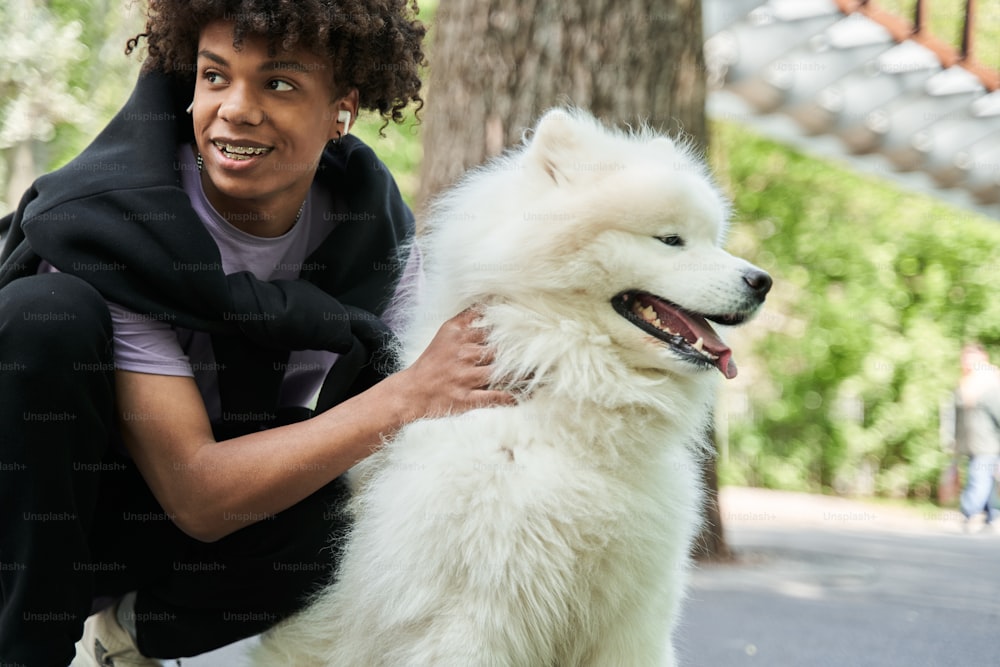 Full length view of the young teen man smiling toothy and looking away while spending time with his pedigree dog in the summer street. Owners and dog care concept