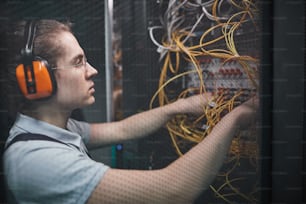 Vista lateral del joven ingeniero de redes que conecta cables en la sala de servidores durante el trabajo de mantenimiento en el centro de datos, espacio de copia