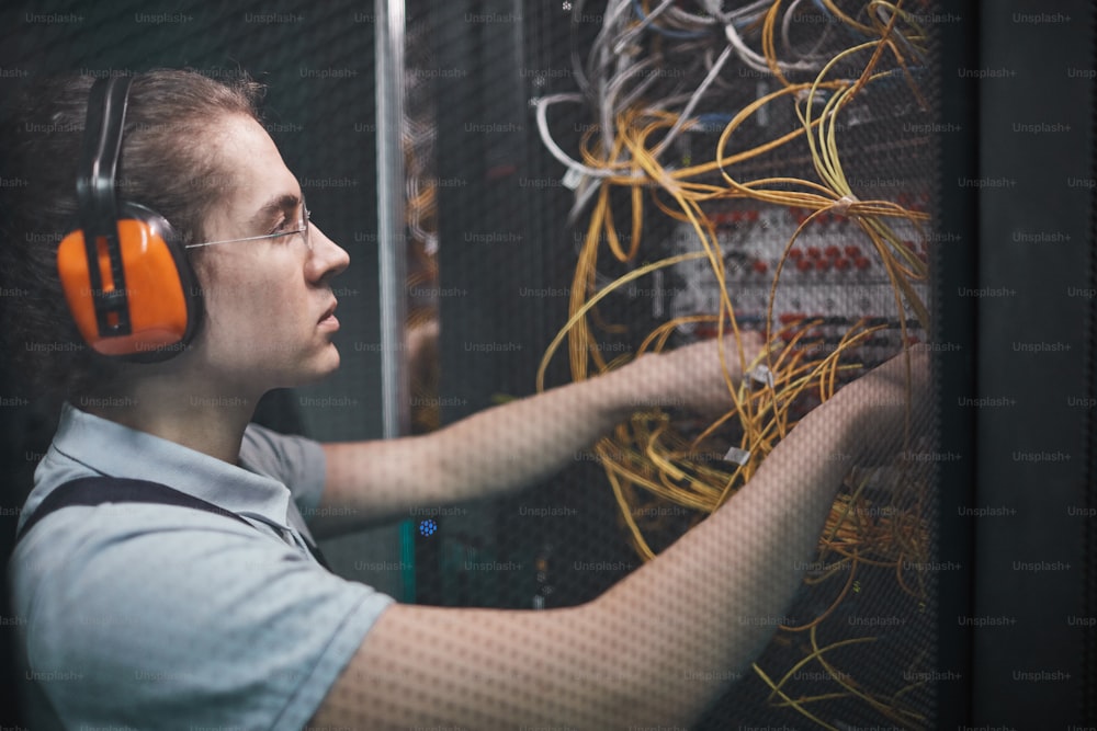 Side view of young network engineer connecting cables in server room during maintenance work in data center, copy space