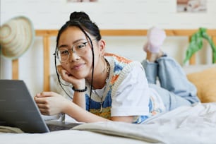 Retrato de una adolescente asiática usando una computadora portátil en la cama en el acogedor interior de la habitación y mirando la cámara, copiar el espacio