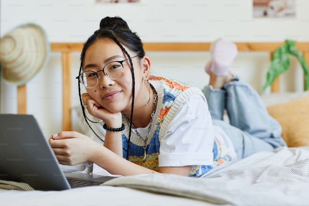 Portrait of teenage Asian girl using laptop on bed in cozy room interior and looking at camera, copy space