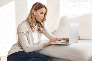 Image of a beautiful blonde woman posing sitting indoors at home using laptop computer.