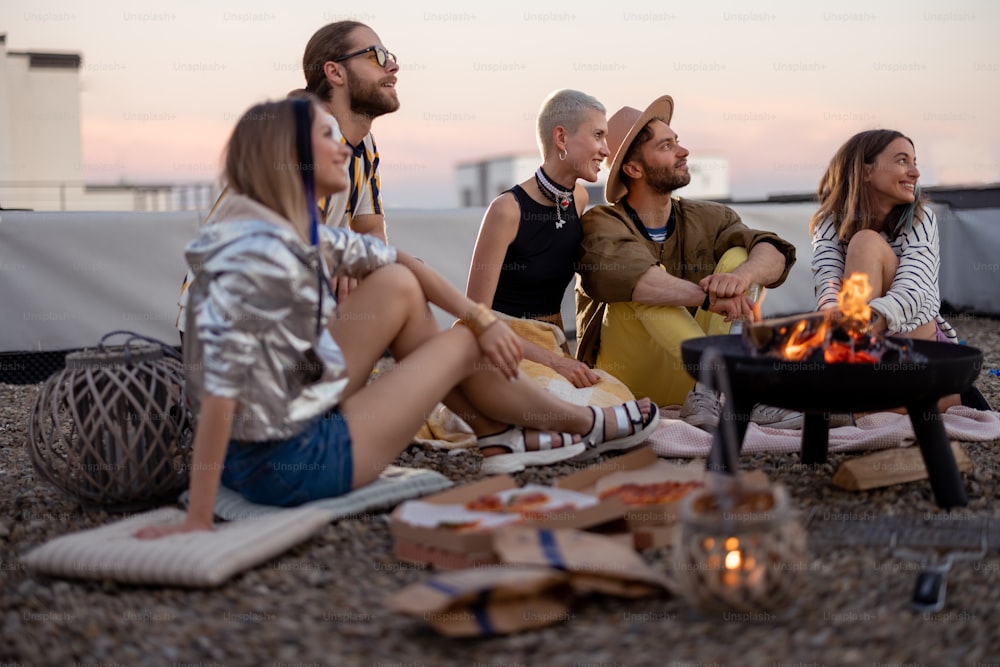 Group of young stylish friends sitting together by the fireplace, having a picnic on the rooftop terrace at dusk. Enjoy summertime and beautiful sunset view