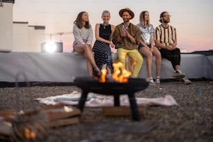 Group of young stylish friends watching cinema, sitting together near the fireplace on the rooftop terrace at dusk. Enjoy summertime and movie outdoors
