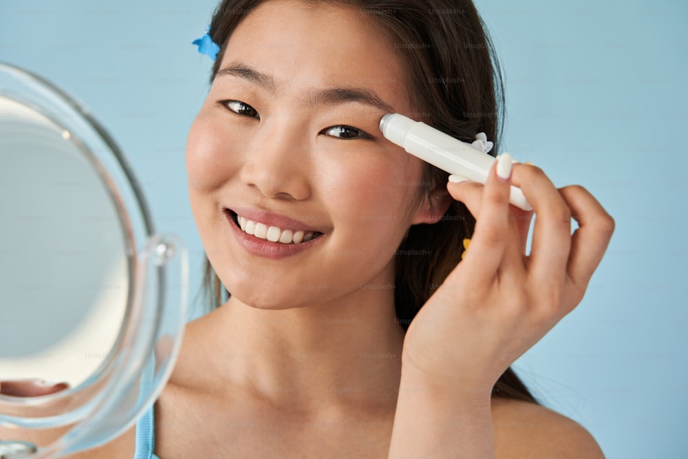 Portrait view of the asian girl with hand mirror looking at the camera while massaging her skin near the eyes with special roller. Stock photo