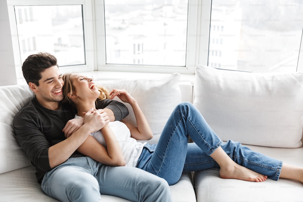Image of smiling couple man and woman hugging together while sitting on sofa in bright room at home