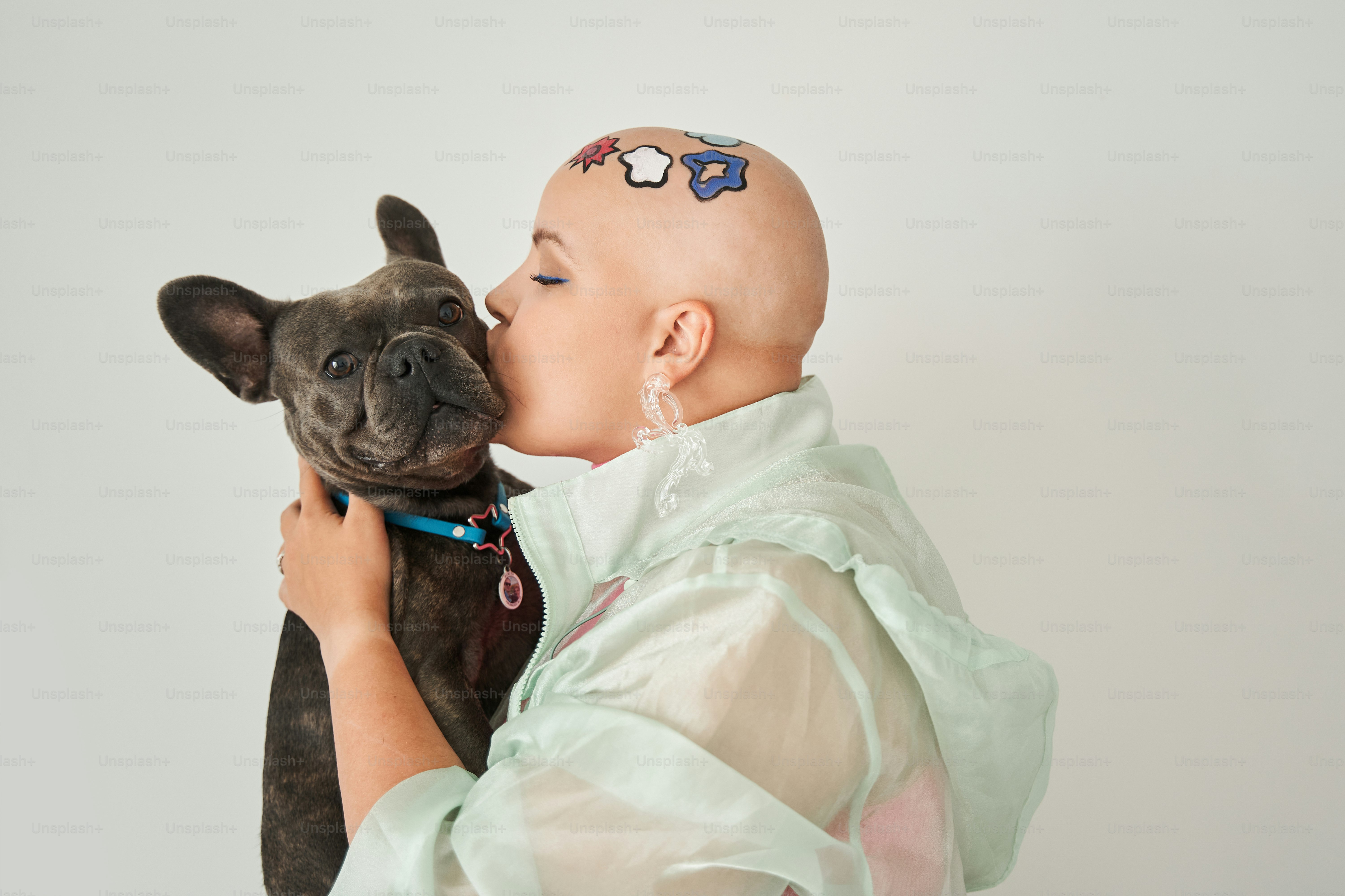 I love you. Portrait view of the bald woman with creative pattern at her head kissing at the muzzle her lovely french bulldog while posing to the camera behind the white wall