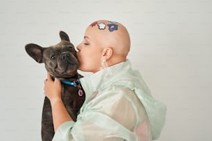 I love you. Portrait view of the bald woman with creative pattern at her head kissing at the muzzle her lovely french bulldog while posing to the camera behind the white wall