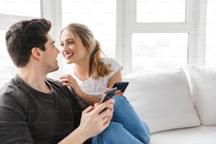 Photo of happy couple man and woman using smartphones together while sitting on sofa in bright room at home