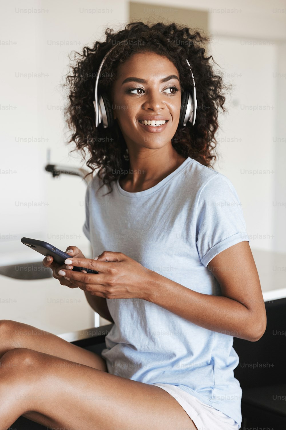 집에서 의자에 앉아 있는 동안 휴대전화를 사용하는 헤드폰을 쓴 매력적인 아프리카 여성
