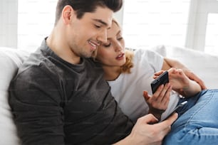 Foto de adorável casal homem e mulher usando smartphones juntos enquanto sentados no sofá em sala brilhante em casa