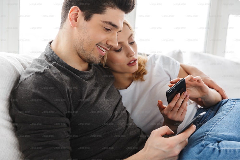 Photo of lovely couple man and woman using smartphones together while sitting on sofa in bright room at home