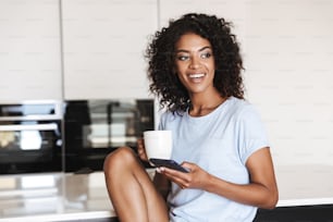Alegre mujer africana usando el teléfono móvil mientras está sentada en una silla en casa con una taza de café