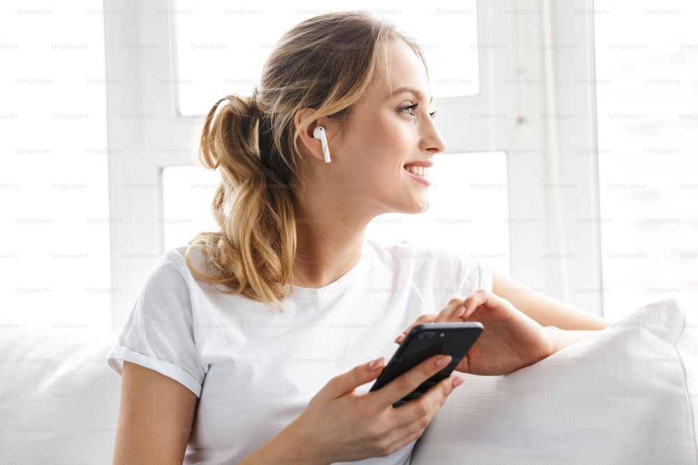 Retrato de una chica caucásica de 20 años en camiseta casual con auricular usando el teléfono móvil mientras está sentada en el sofá en una habitación luminosa en casa