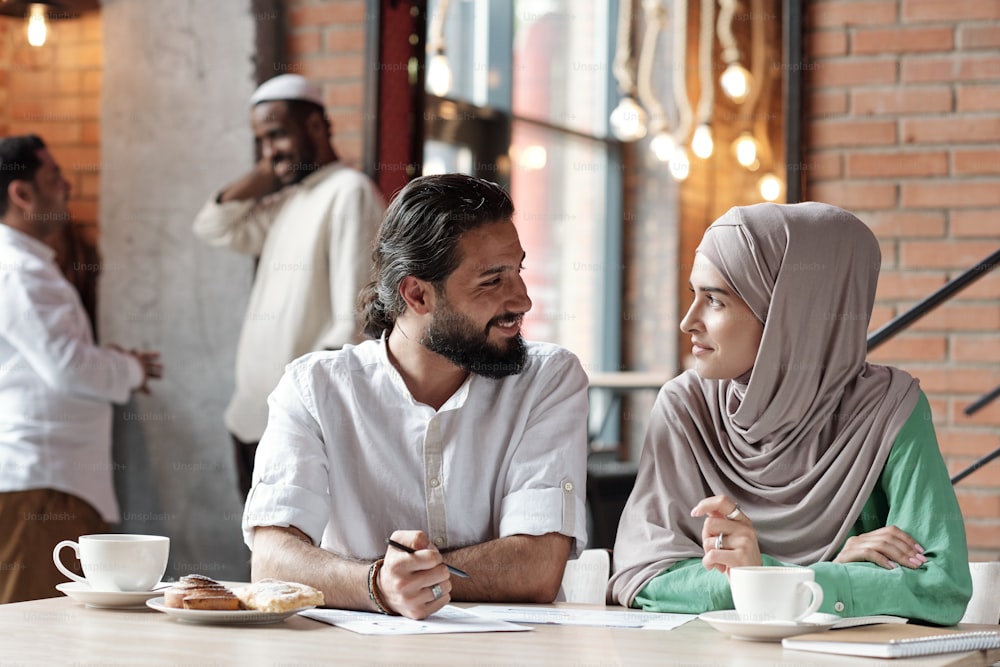 Homme d’affaires musulman barbu souriant discutant de papiers avec une femme en hijab alors qu’ils travaillent dans un café