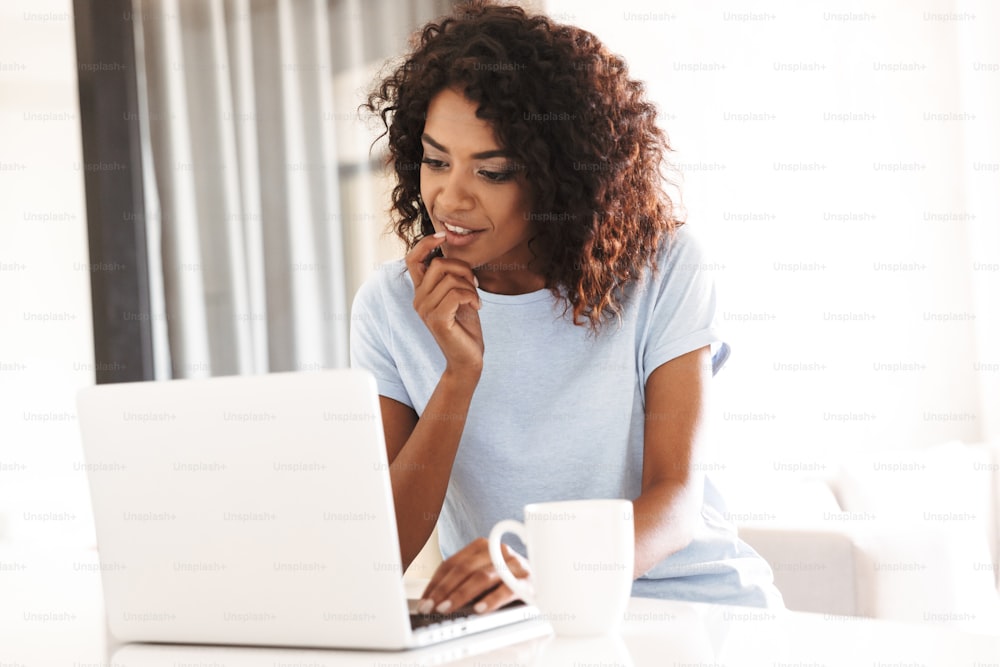 Mujer africana pensativa que usa una computadora portátil mientras está sentada en casa con una taza de café