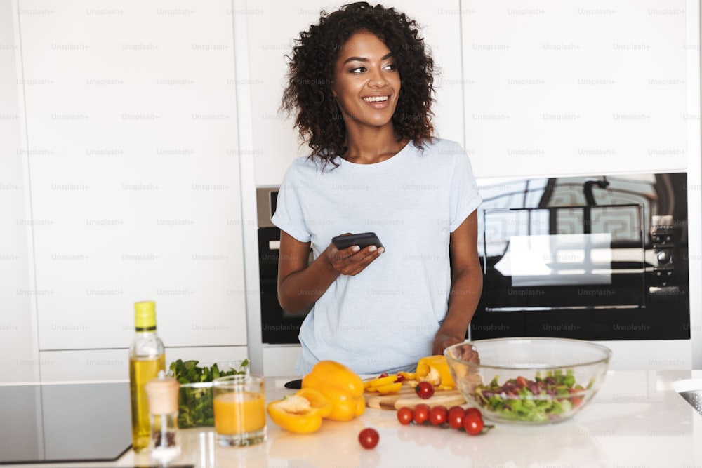 Ritratto di una donna afroamericana sorridente che usa il telefono cellulare mentre prepara un'insalata sana in cucina