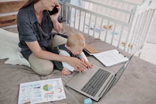 Multi-tasking young mother sitting with baby on bed and answering phone call while baby reaching hands to computer