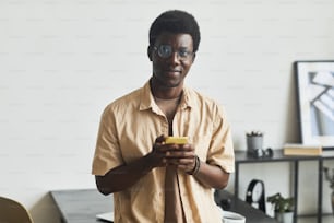 Portrait d’un jeune homme africain regardant un appareil photo et utilisant son téléphone portable alors qu’il se tenait au bureau