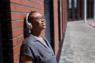 African happy woman in wireless headphones leaning on the brick wall enjoying the music outdoors