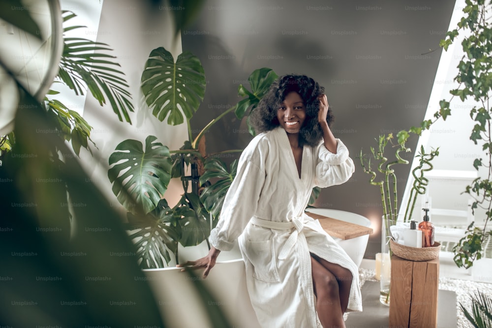 Time for yourself. Smiling dark-skinned pretty long-haired woman in light robe sitting on bathtub surrounded by green plants