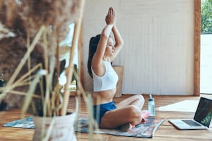 Belle jeune femme en tenue de sport pratiquant le yoga sur la terrasse avec un ordinateur portable allongé près d’elle