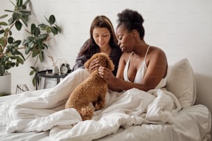 Two happy young women cuddling cute dog while relaxing in bed