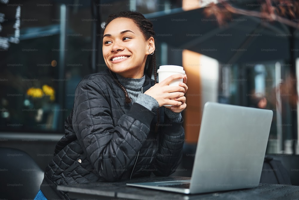 Smiling young woman expressing happiness whiles sitting at the table with a laptop and holding a paper cup of coffee