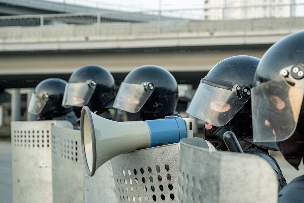 Un policier portant un casque anti-émeute debout parmi des collègues avec des boucliers anti-émeute et parlant en méfaphone tout en neutralisant les terroristes