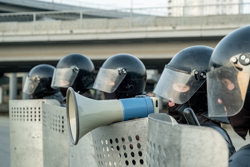 Policial com capacete antimotim em pé entre colegas de trabalho com escudos antimotim e falando em mefaphone enquanto neutraliza terroristas