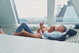 Beautiful mature couple examining smart phone and smiling while relaxing in big hammock at home together