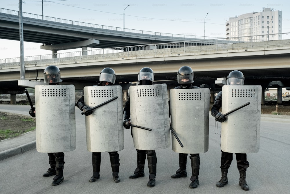 Grupo de guardias policiales uniformados protegidos por escudos de pie con asas laterales y escudos contra puentes de la ciudad