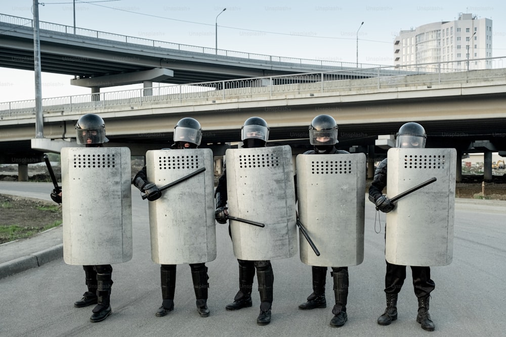 Group of police guards in uniform protected by shields standing with side handles and shields against city bridges