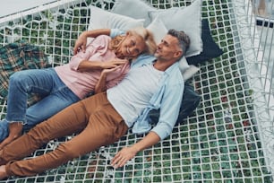 Top view of beautiful mature couple smiling and communicating while relaxing in big hammock at home together
