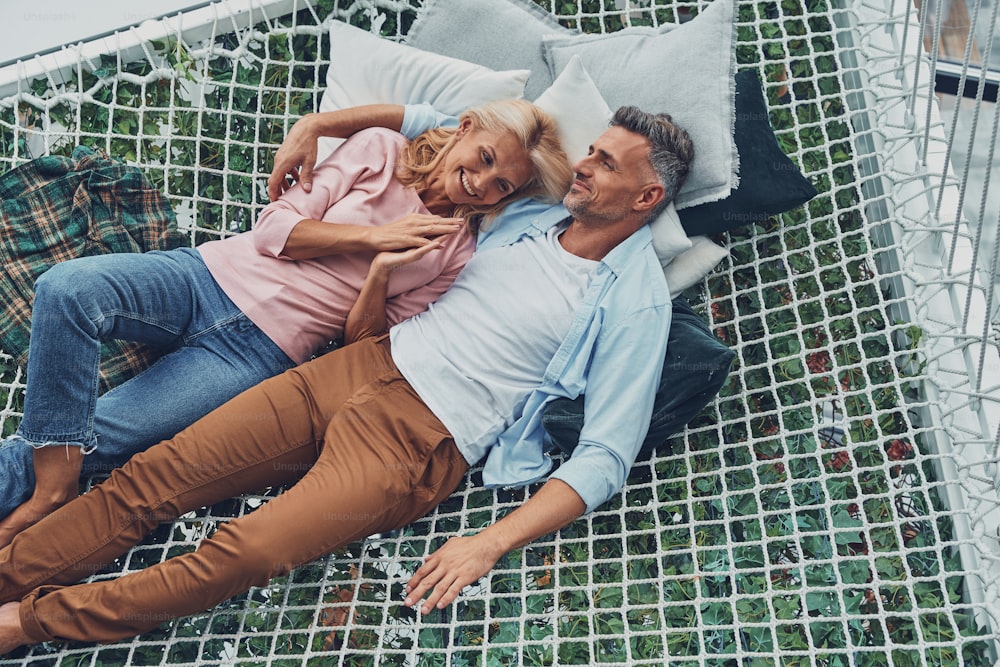 Vue de dessus d’un beau couple d’âge mûr souriant et communiquant tout en se relaxant dans un grand hamac à la maison ensemble
