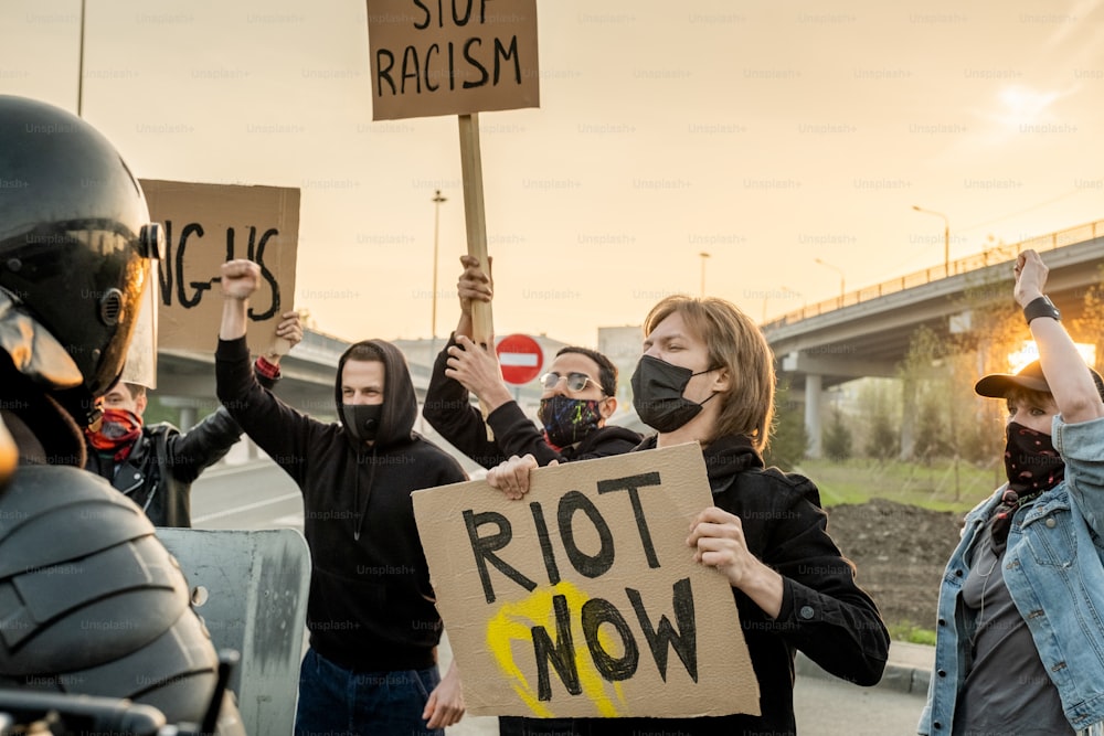 Gruppo di persone scontente in maschera con cartelli che rivendicano uguali diritti per tutte le etnie mentre urlano contro la polizia antisommossa sulla strada