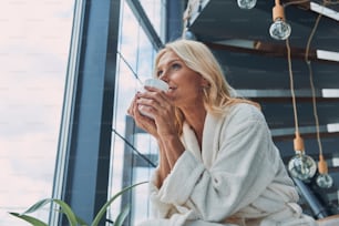 Beautiful mature woman in bathrobes drinking coffee and smiling while standing near window at home