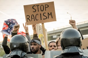 Grupo de jovens americanos desagradados ergue punhos e bandeira enquanto pede para parar o racismo, polícia mantém multidão