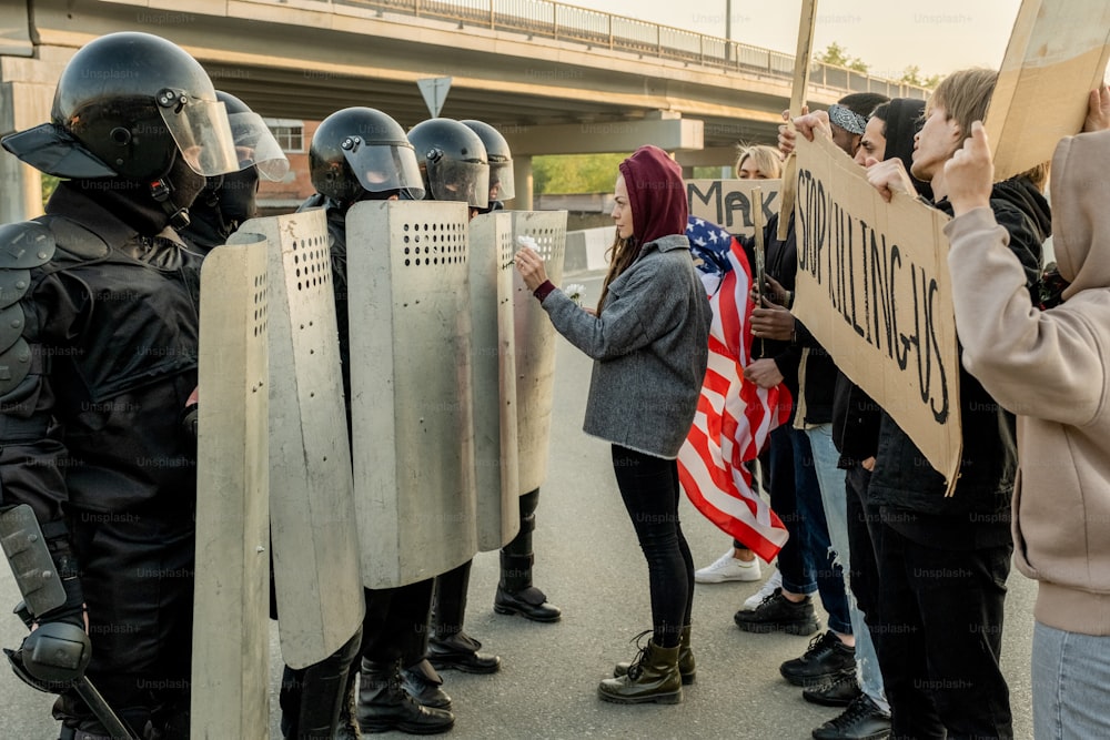 Young woman in hood standing in front of police guards and giving daisy to them while trying to stop violence between police and protestors