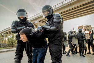 Riot police in body armors twisting man into uncomfortable position while confronting crowd in city