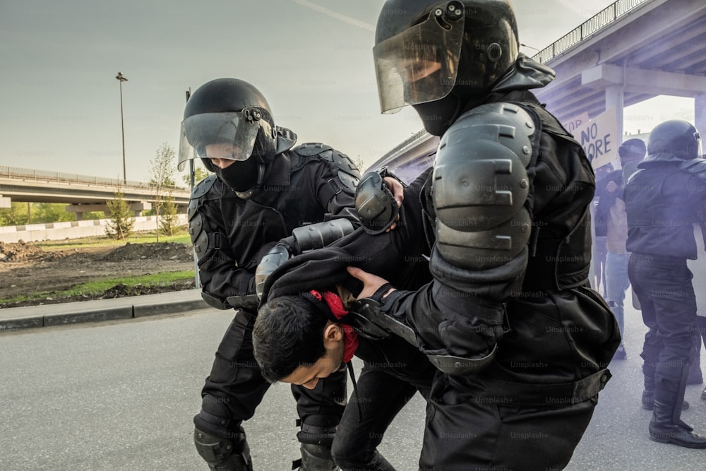 Erfahrene Bereitschaftspolizisten mit Helmen, die die Hände des Rebellen hinter seinem Rücken halten, während sie Menschen bei einer Kundgebung verhaften