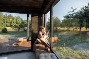 Mujer joven descansando en una hermosa casa de campo u hotel, sentada en el alféizar de la ventana disfrutando de una hermosa vista sobre el bosque de pinos y haciendo una foto por teléfono. Concepto de soledad y recreación en la naturaleza